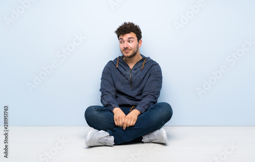 Young man sitting on the floor making doubts gesture looking side © luismolinero