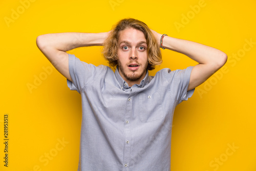 Blonde man over isolated yellow background frustrated and takes hands on head