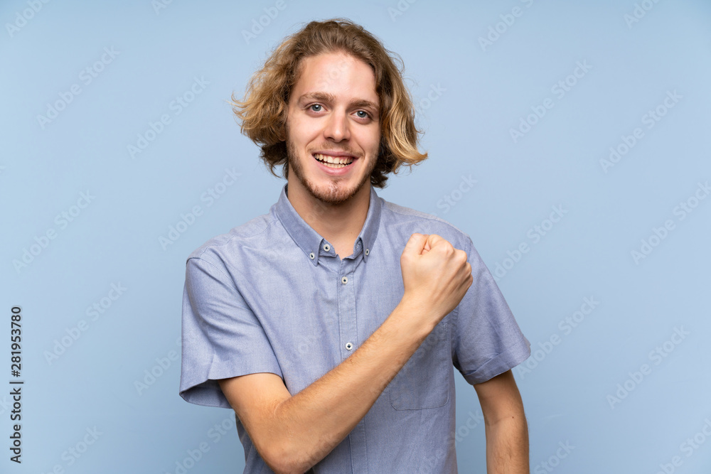 Blonde man over isolated blue wall celebrating a victory Blonde man over isolated blue wall celebrating a victory