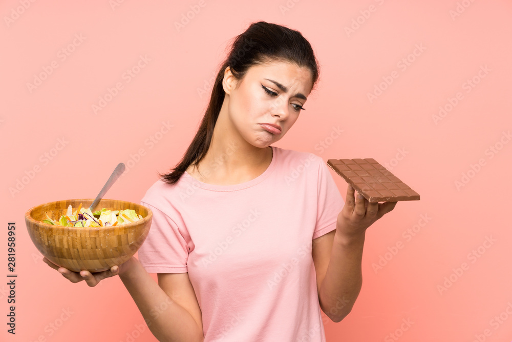 Teenager girl over isolated pink wall with salad and chocolat and having doubts Teenager girl over isolated pink wall with salad and chocolat and having doubts