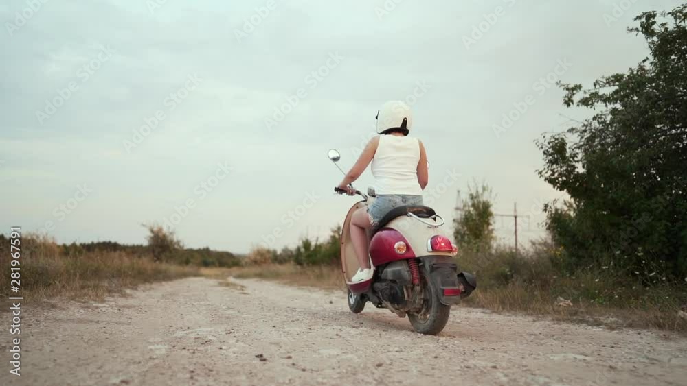 Woman in helmet rides on retro moped along country road in summer at sunset. Camera shoots from behind Woman in helmet rides on retro moped along country road in summer at sunset. Camera shoots from behind