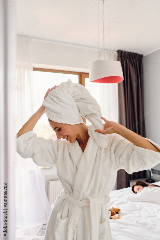 Young cheerful woman in white bathrobe and towel on head happily looking down while pretty girl friend lying in bed on background in modern hotel