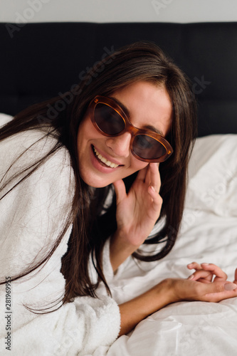 Portrait of young beautiful woman with dark hair in white bathrobe and sunglasses happily lying on big bed in modern hotel