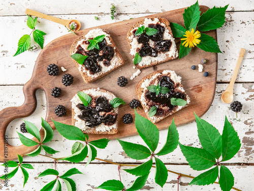 Mini sandwiches with black bread, cottage cheese and jam of berries on rustic cutting board on a white wooden table. Top view