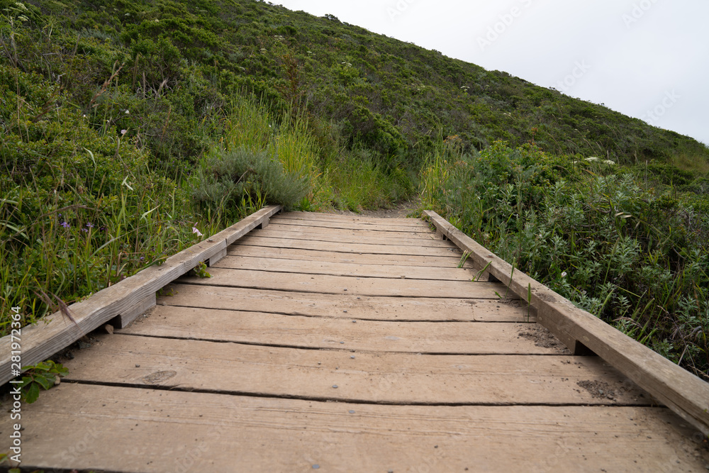 Small wooden bridge leading on hiking trail connecting grassy hills