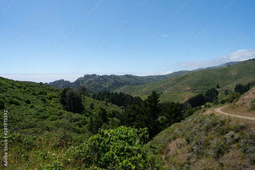 Blue skies, grassy hills, man on hiking path, city in background Blue skies, grassy hills, man on hiking path, city in background