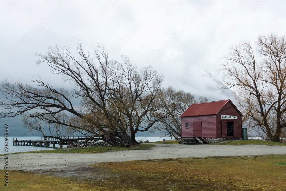 Red wooden shed on the bank of the Wakatipu lake with mountains on the background at Glenorchy village during foggy day. New Zealand.