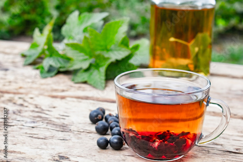 Tea in a glass cup of black currant on a wooden table near a glass teapot on a background of green grass.