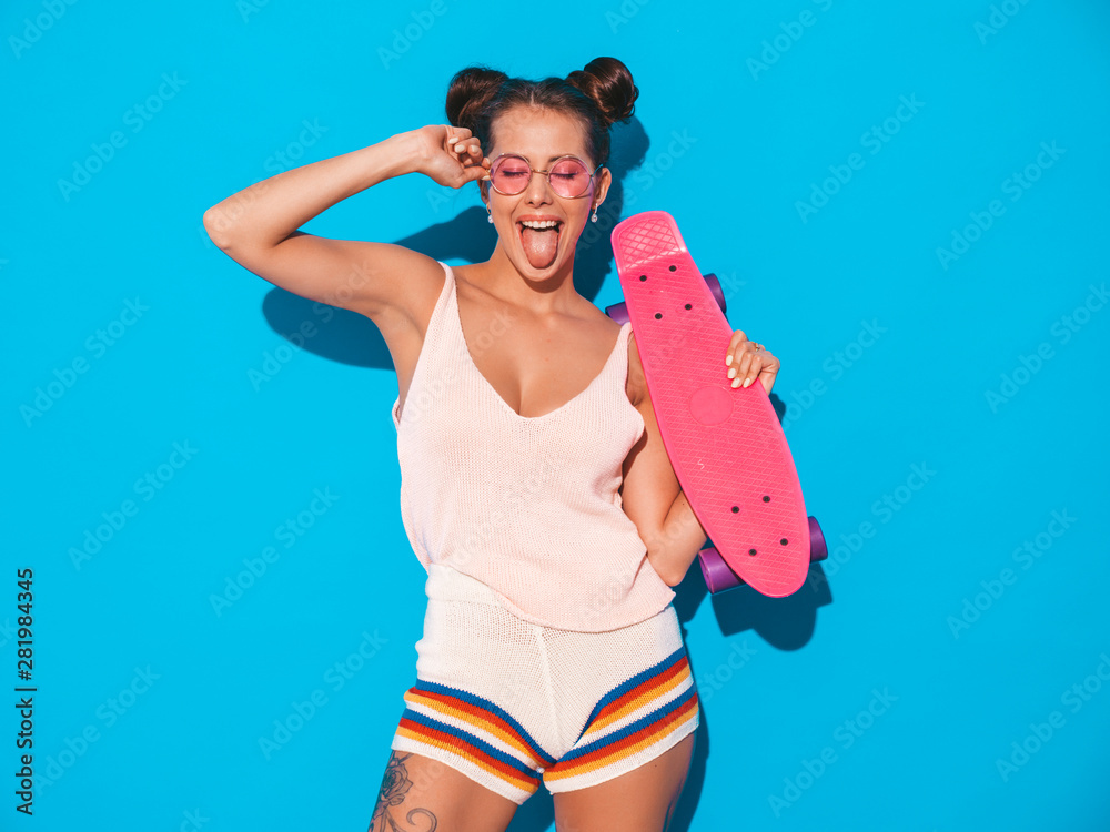 Young beautiful sexy smiling hipster woman in sunglasses.Trendy girl in  summer knitted cardigan topic ,shorts.Positive female going crazy with pink  penny skateboard,isolated on .blue wall.Two horns Stock Photo | Adobe Stock