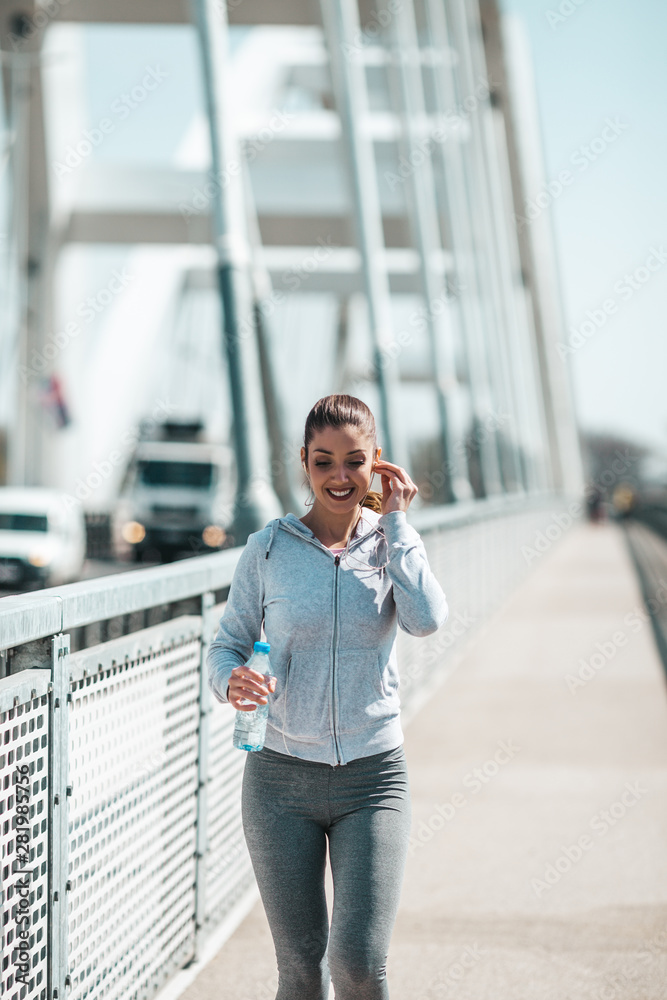 Fototapeta premium Beautiful young and fit woman in good shape running and jogging alone on city bridge street. She wears sporty earbuds and listens to music.