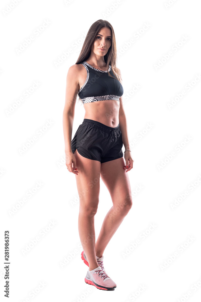 Attractive young fitness woman in black top and shorts posing at camera with blank expression. Full body isolated on white background. 