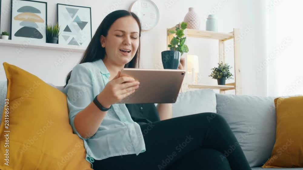 Young woman seat on sofa with tablet and drink coffe or tea from blue cup in living room. Slow motion 4k footage. Young woman seat on sofa with tablet and drink coffe or tea from blue cup in living room. Slow motion 4k footage.