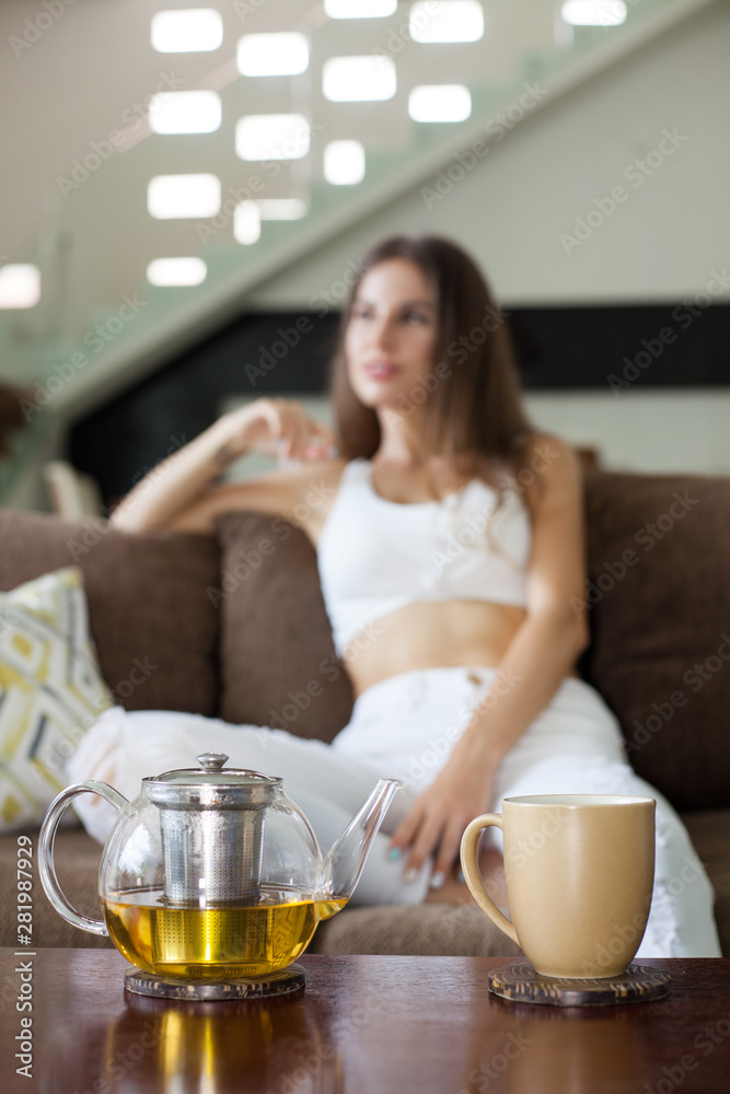 vertical. glass teapot with herbal tea and mug on the table. unfocused beautiful smiling girl sitting on the sofa in the background vertical. glass teapot with herbal tea and mug on the table. unfocused beautiful smiling girl sitting on the sofa in the background