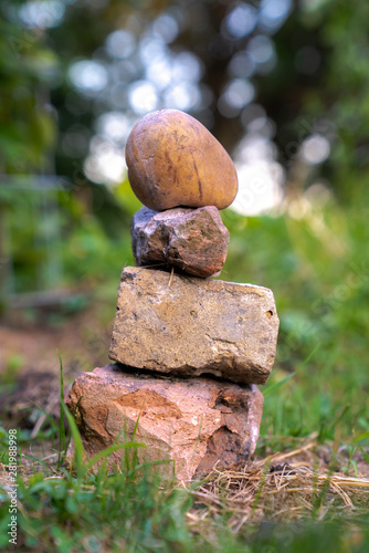 A stack or pile of small rocks with earth tone colors layered on one another and sitting in the grass near a garden with blurred bokeh background with sun light shining through the trees beyond.