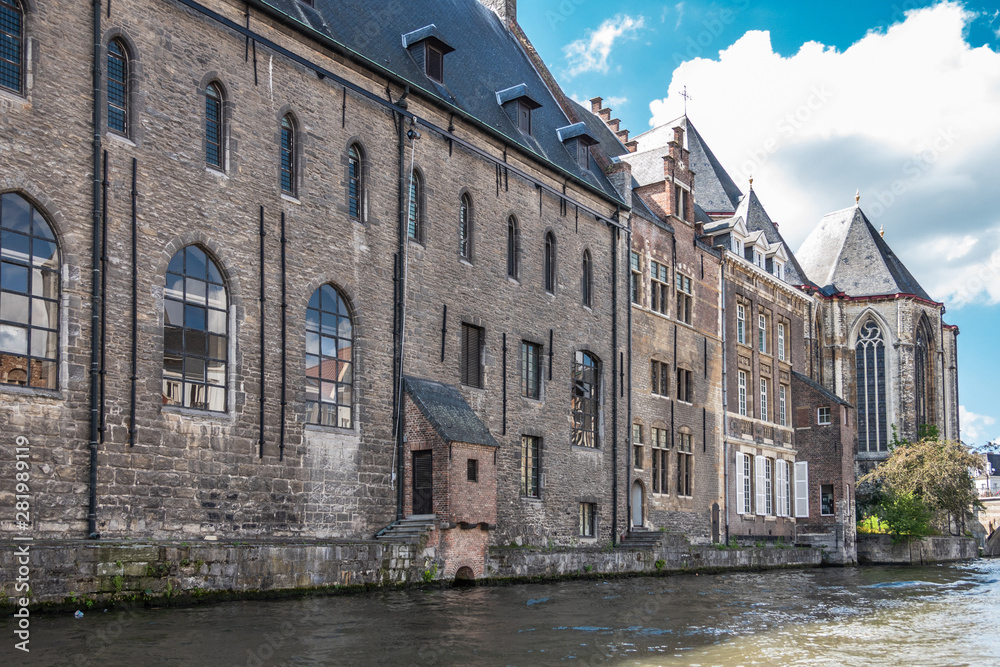 Gent, Flanders, Belgium -  June 21, 2019: Brown stone University building Het Pand bordering Leie River under blue-white cloudscape. Greenish water, Green foliage.