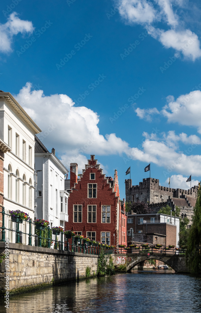 Gent, Flanders, Belgium -  June 21, 2019: Narrow Lieve River leads along old bourgeois mansions to Gravensteen Castle under blue sky with white clouds. Bluish water. Flowers on quays.