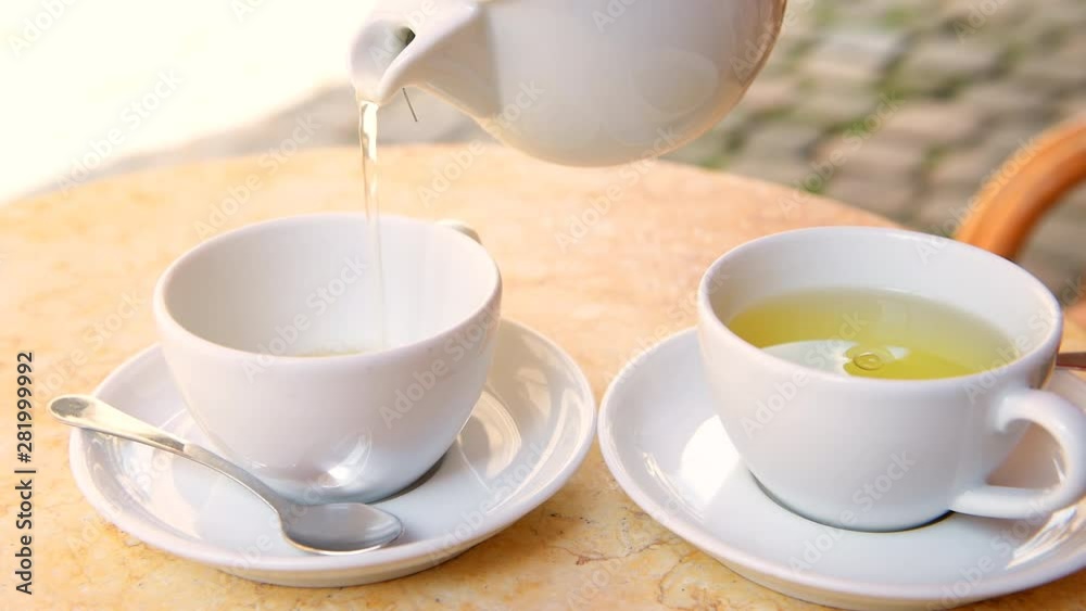 Closeup of young woman sitting at summer restaurant or cafe table, hands holding and pouring hot green tea in cup from teapot in city of Lviv or Lvov, Ukraine Closeup of young woman sitting at summer restaurant or cafe table, hands holding and pouring hot green tea in cup from teapot in city of Lviv or Lvov, Ukraine