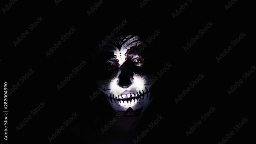 Halloween mask. A girl in a blinking and frightening light. Mexican day of the dead. Portrait of a young woman with scary makeup for Halloween on a dark background. 4K