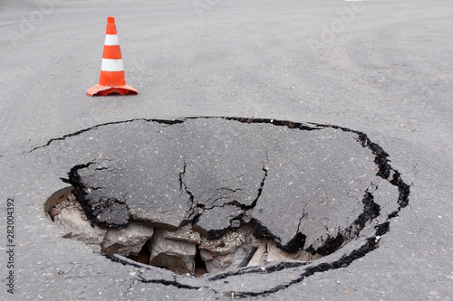 Deep sinkhole on a street city and orange traffic cone. Dangerous hole in the asphalt highway. Road with cracks. Bad construction. Damaged asphalt road collapse and fallen. photo