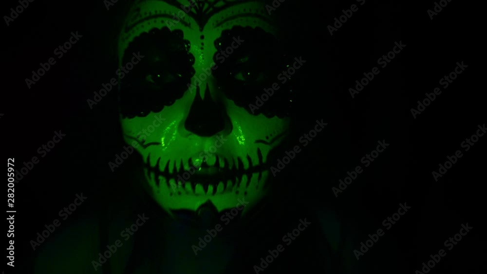 Halloween mask CALAVERA CATRINA. A girl in a blinking and frightening light. Mexican day of the dead. Portrait of a young woman with scary multi-colored makeup for Halloween on a dark background. 4K Halloween mask CALAVERA CATRINA. A girl in a blinking and frightening light. Mexican day of the dead. Portrait of a young woman with scary multi-colored makeup for Halloween on a dark background. 4K