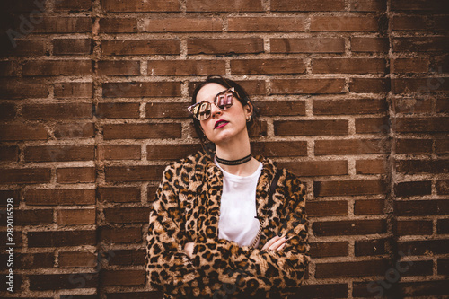 Stylish portrait of young serious woman with crossed arms wearing trendy leopartd print faux fur coat, fashion sunglasses, posing on red brick wall texture grunge background