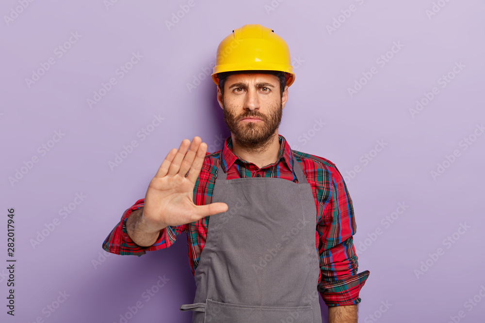 Displeased male engineer makes rejection gesture, says no, keeps palm outstretched at camera, refuses doing manual work, wears hardhat, casual uniform, isolated on purple wall. Serious constructor