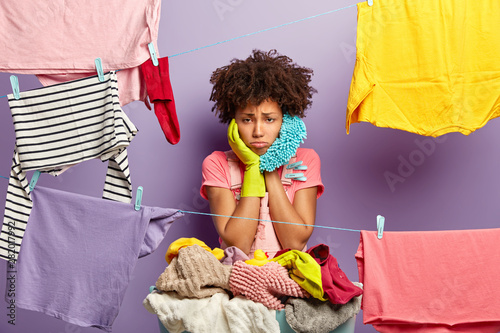 Upset overworked housewife hangs clothes on washing line with clothespins, busy doing housework, wears casual outfit and rubber protective gloves, surrounded with laundry. Washing day concept photo
