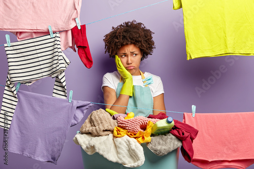 Laundry day concept. Fatigue dissatisfied woman touches cheek and looks away in unhappiness, stands near basket with pile of laundry and detergent, washing lines with hanged wet clothes for drying photo