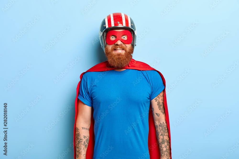 Glad bearded man with funny outlook, comes on costume party, being superhero  character, wears helmet, mask and red cloak, has fun with friends, poses  against blue background. Save world concept Stock Photo |