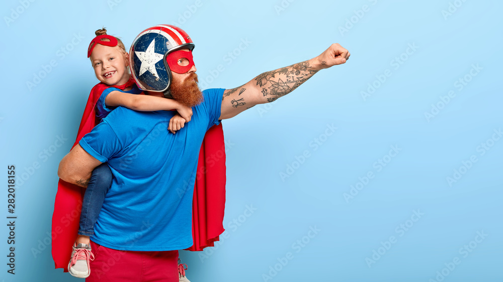 Indoor shot of powerful dad in protective helmet carries small child on back, play superhero game, fly together and make good things for people, isolated on blue background. Childhood, parents concept Indoor shot of powerful dad in protective helmet carries small child on back, play superhero game, fly together and make good things for people, isolated on blue background. Childhood, parents concept