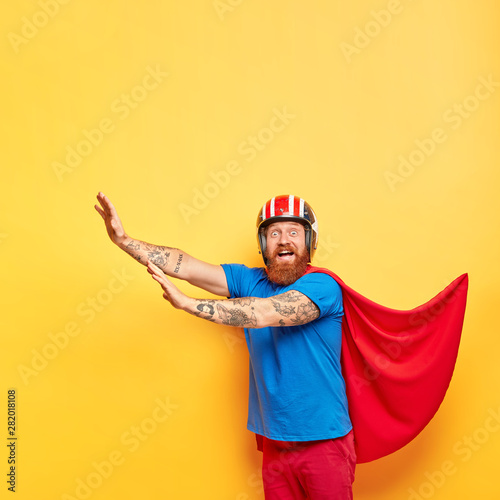 Image of surprised glad bearded guy wears special costume, comes on party, makes defense gesture, isolated on yellow background. Cheerful superhero in helmet, cape, imagines fight, feels brave