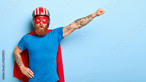 Stupefied emotive man with ginger beard being cartoon character, keeps arm in flying gesture, wears protective headgear, blue t shirt and red cloak, has shocked expression, saves our universe photo