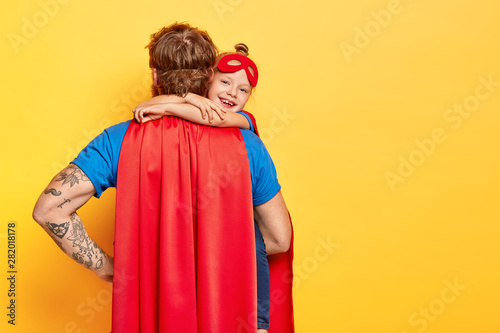 Lovely glad kid embraces tightly father who stands back to camera, wears red cloak, plays together with daughter, pretend being superhero, isolated on yellow background with empty space for text