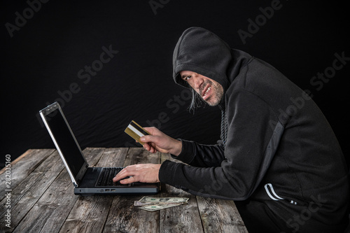 hacker in black hoody with laptop, bank card and dollar notes