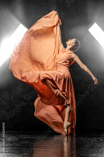 Ballerina. A young dancer dressed in a long peach dress, pointe shoes with ribbons. Performs a graceful, graceful dance movement on the background of the bright light of the window.  Ballet studio. 