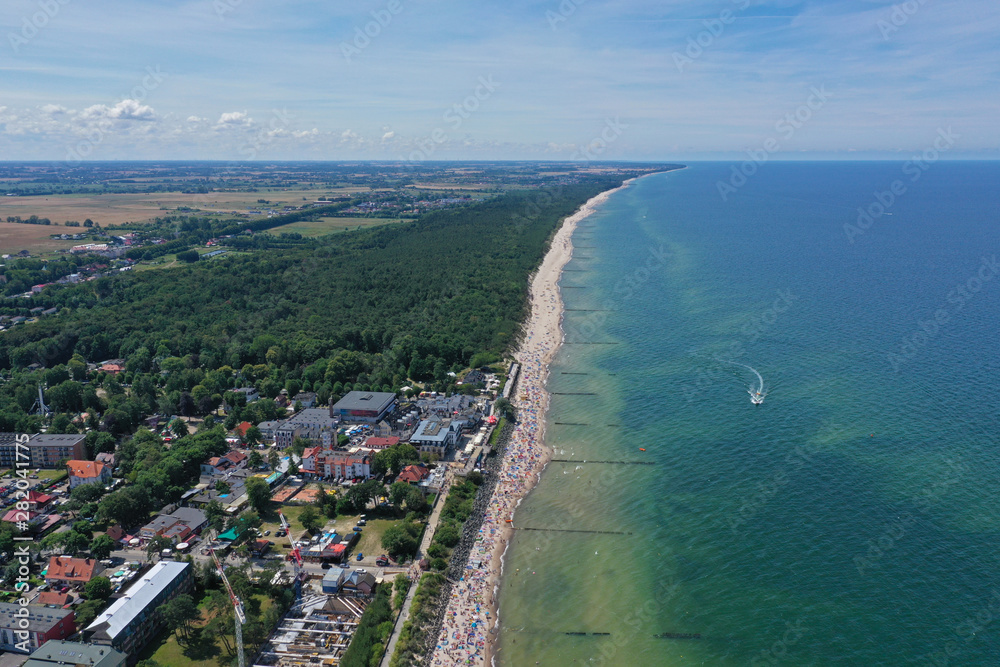 Drone aerial perspective view on sunny beach with sunbathers with windbreaks and towels at sea in touristic city.