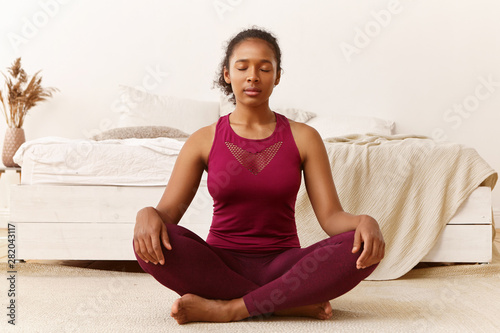 Meditation, peace, balance, zen and harmony concept. Portrait of healthy fit young African woman with eyes closed sitting on floor after yoga practice, meditating, doing breathing exercises © Anatoliy Karlyuk