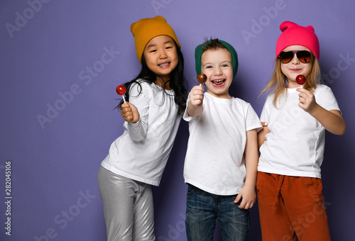 Company of stylish joyful kids in white t-shirts and colorful hats enjoy sweet lollipops candies the have got