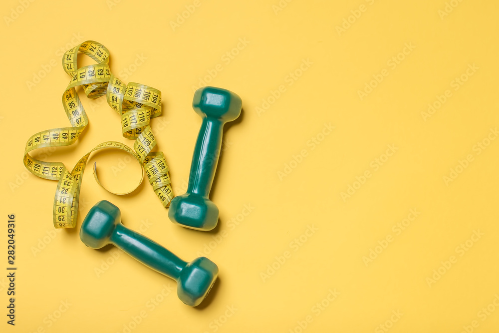 Dumbbells and measuring tape on yellow background, top view