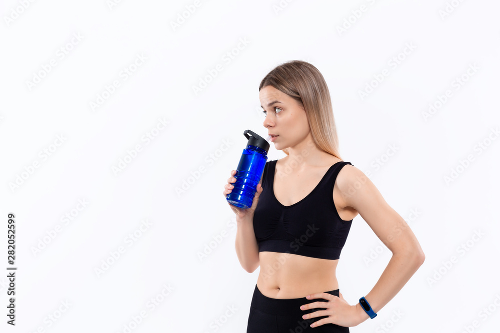 Young sporty blond woman in a black sportswear with smart watches drinks water after workout standing over white background. Young sporty blond woman in a black sportswear with smart watches drinks water after workout standing over white background.