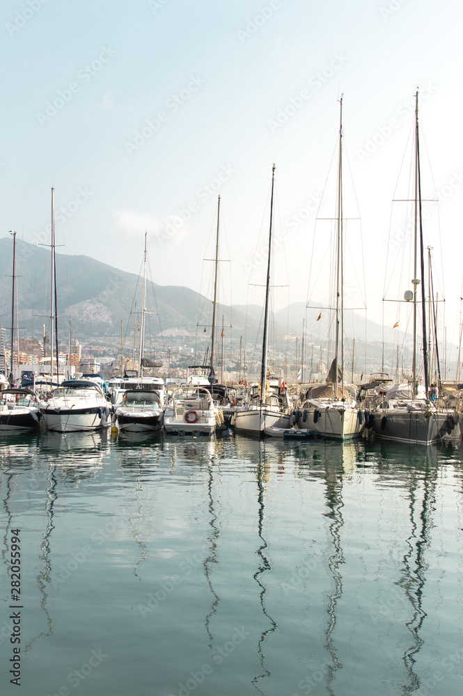 Sailboats and yachts in the dock reflected in the water with the city and mountains in the background. Landscape of Costa del Sol (Fuengirola), Spain.
