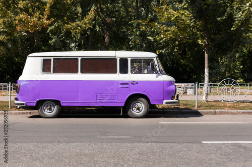 purple Retro van standing on the road near the fence, a park in the background. Classic wagon car is ready for summer vacations and trips to the sea