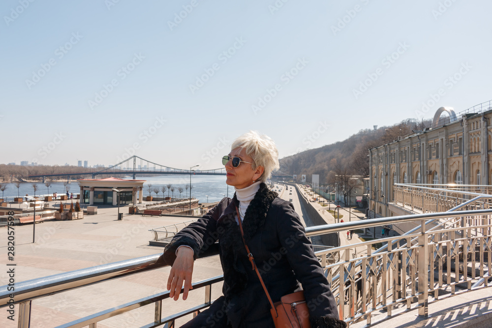 fashionable adult european woman in black glasses posing at the rail against the background of the highway and the bridge across the river. middle-aged woman tourist and traveler.