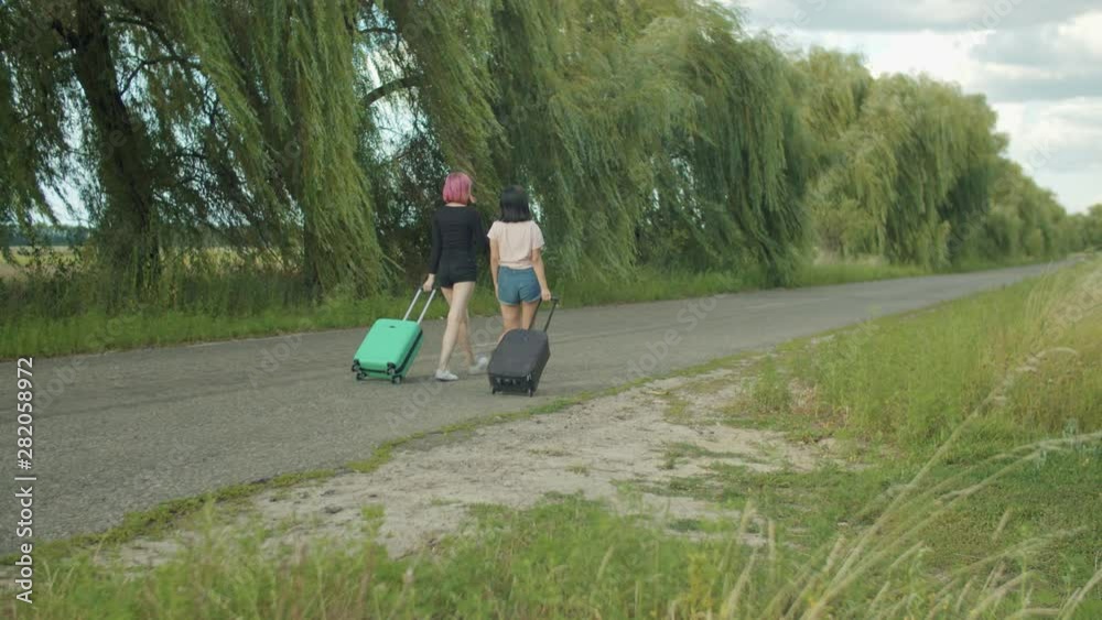 Rear view of chatting carefree diverse female tourists with travel bags walking along rural road while travelling on summer vacations. Independent young women exploring countryside during hiking. Rear view of chatting carefree diverse female tourists with travel bags walking along rural road while travelling on summer vacations. Independent young women exploring countryside during hiking.
