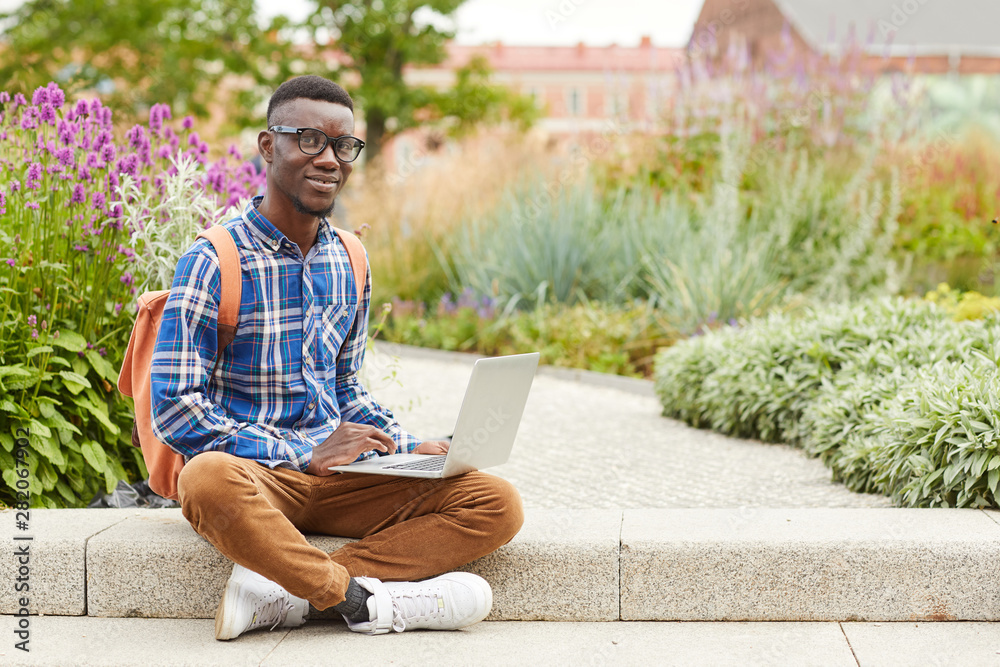 Fototapeta premium Full length portrait of African student smiling at camera while using laptop outdoors in college campus, copy space