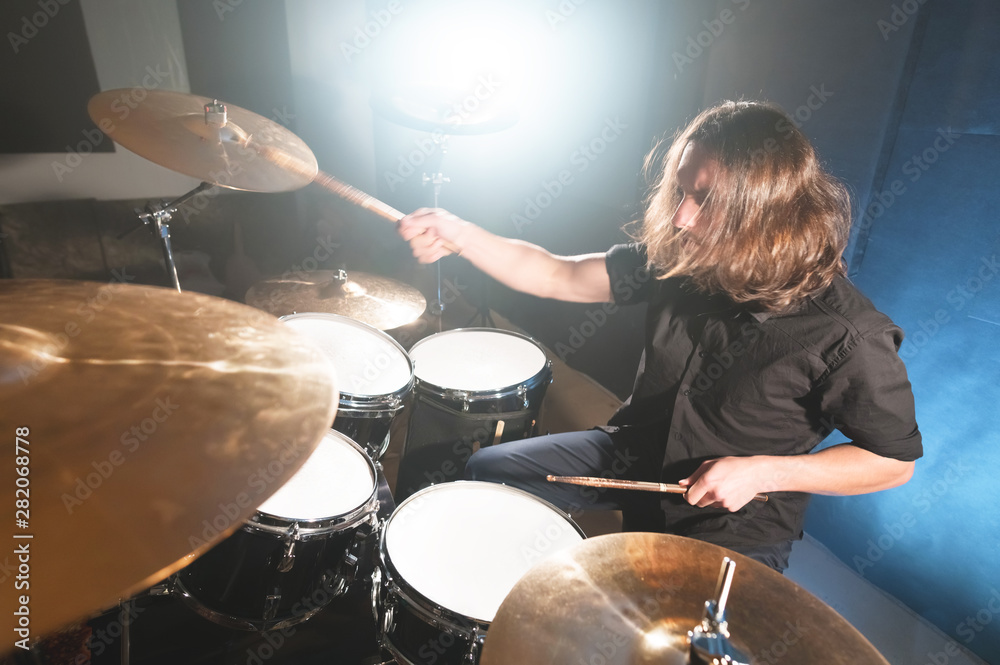 Obraz premium Portrait of a long-haired drummer with chopsticks in his hands sitting behind a drum set. Low key. Concepts of the creative freedom of the millenial generation