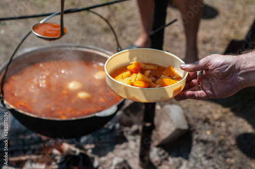 Ukrainian borsch. Traditional red soup in a large metal cauldron on a bonfire. Cooking on a camping trip. Hand holds a plate in which soup is poured.