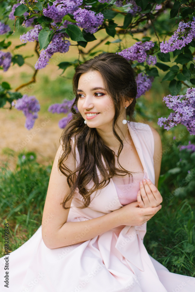 Beautiful young girl sitting on the ground in the garden with blooming lilac. She is happy and smiles a beautiful smile with white teeth. Look away