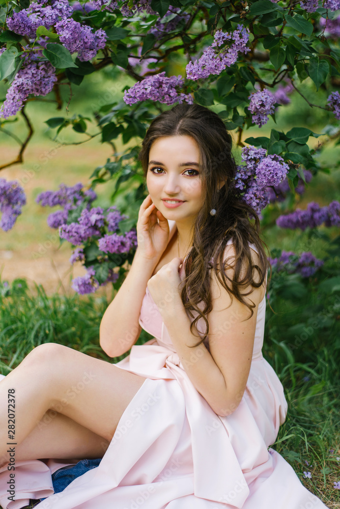 Beautiful young girl sitting on the ground in the garden with blooming lilac. She is happy and enjoys her youth and spring. Professional makeup and clean beautiful skin Beautiful young girl sitting on the ground in the garden with blooming lilac. She is happy and enjoys her youth and spring. Professional makeup and clean beautiful skin