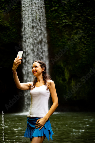 Happy Caucasian woman enjoying waterfall landscape, taking selfie with smartphone near waterfall.